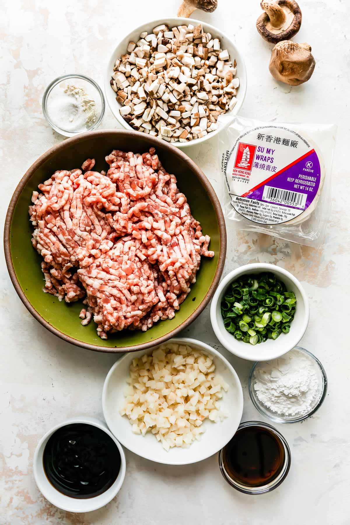 Pork shumai ingredients are arranged atop a creamy white textured surface: ground pork, cornstarch slurry, sesame oil, green onions, dried shiitake mushrooms, water chestnuts, oyster sauce, soy sauce, granulated sugar, white pepper, and round wonton wrappers.