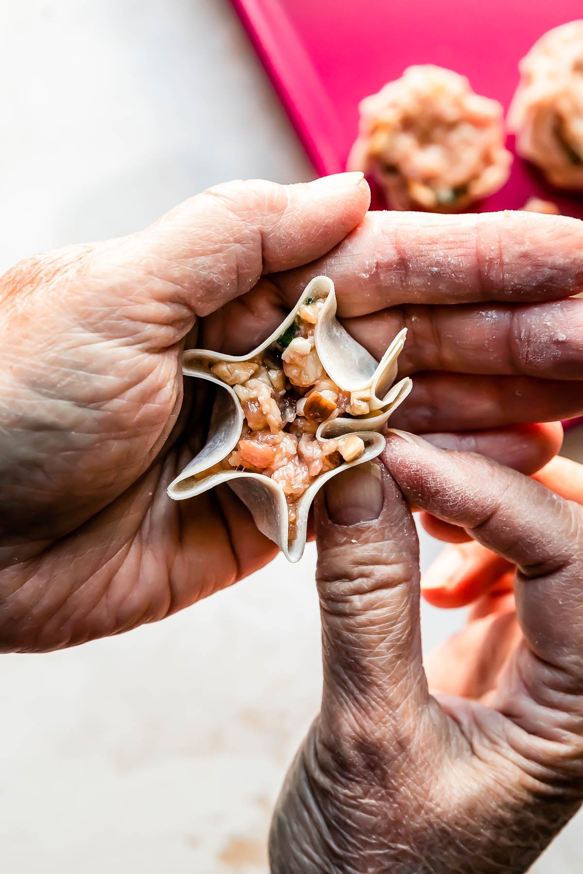 A woman's hands form a shumai dumpling into a purse shape over a creamy white textured surface. A pink baking sheet filled with a few assembled shumai dumplings rests atop the surface.