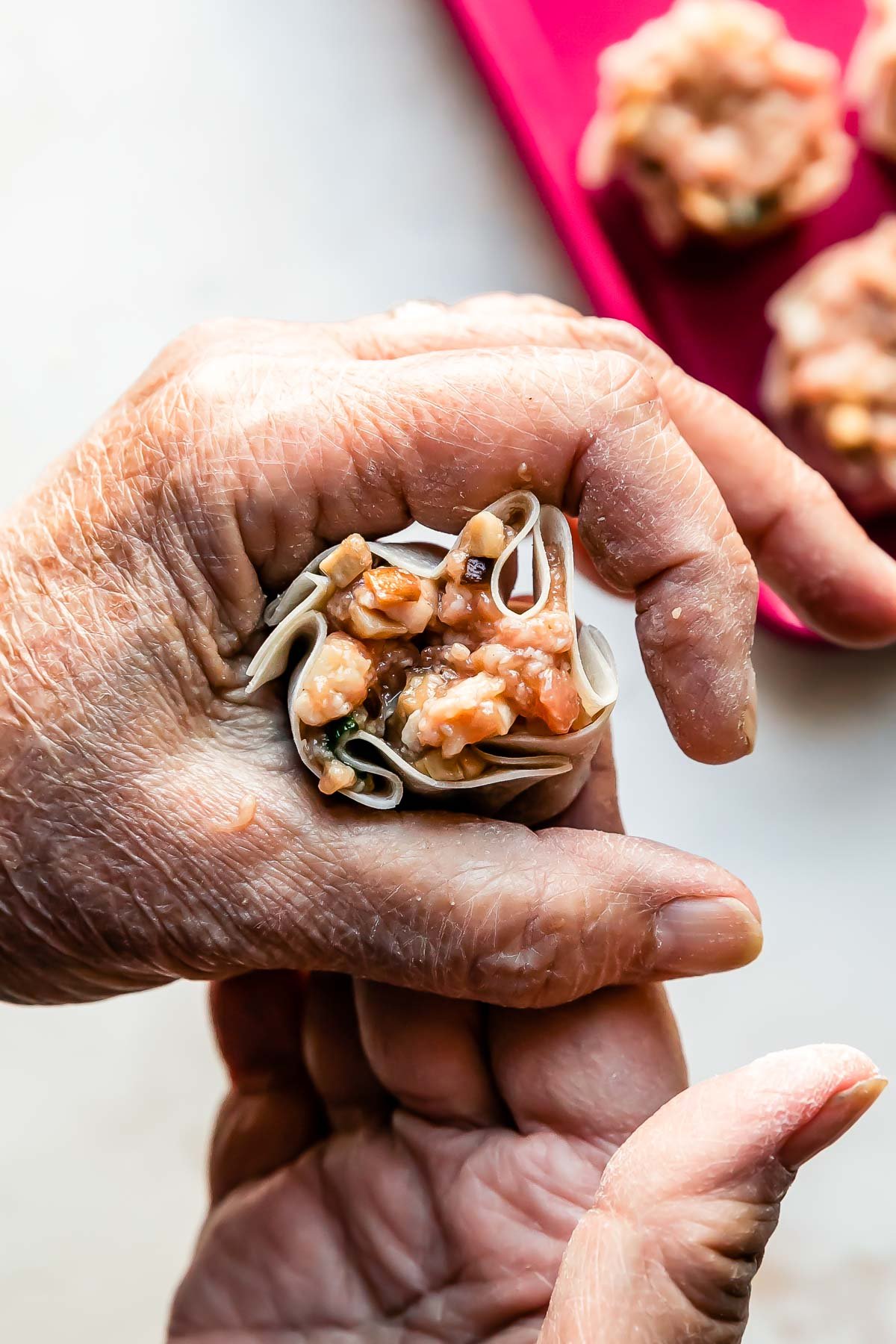 A woman's hands form a shumai dumpling into a purse shape over a creamy white textured surface. A pink baking sheet filled with a few assembled shumai dumplings rests atop the surface.