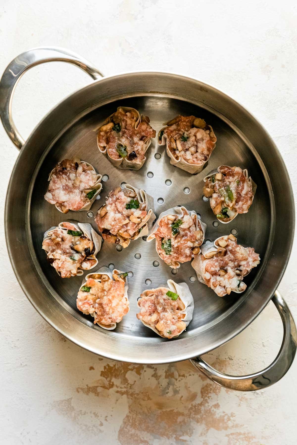 Ten siu mai dumplings rest inside of a metal steamer basket that sits atop a creamy white textured surface.