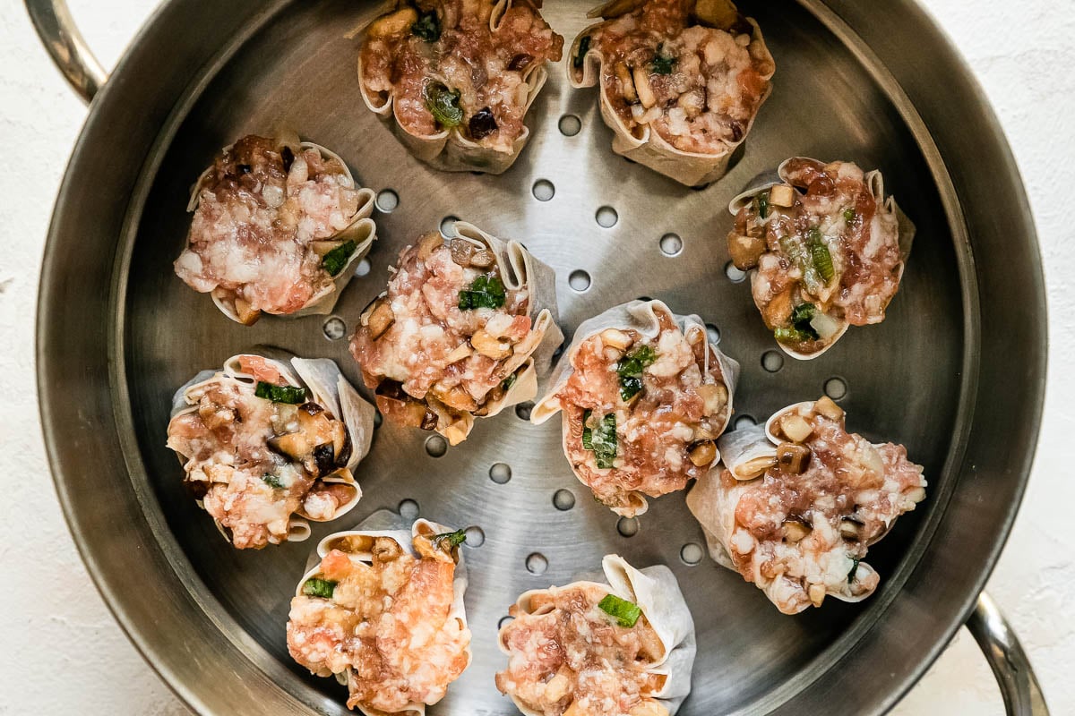 Ten siu mai dumplings rest inside of a metal steamer basket that sits atop a creamy white textured surface.