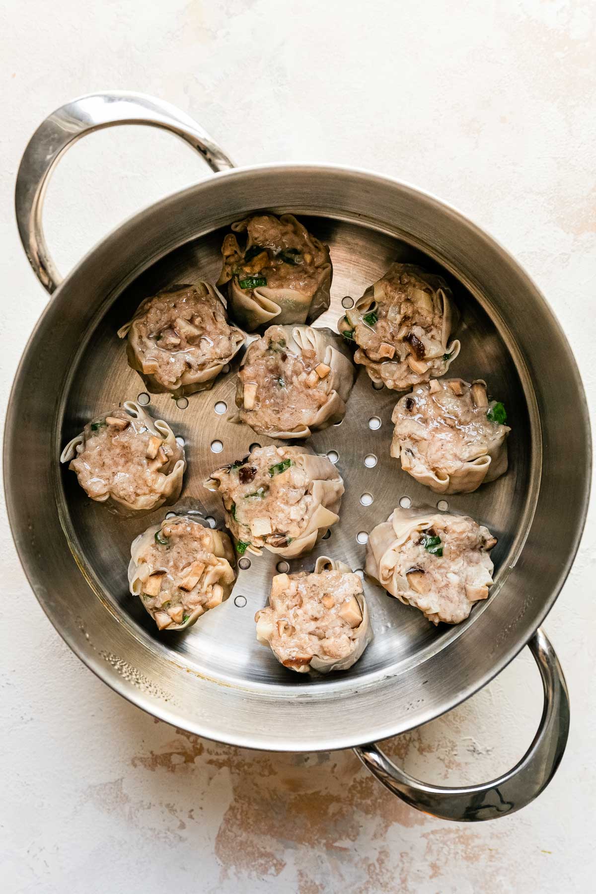 Ten steamed shumai dumplings rest inside of a metal steamer basket that sits atop a creamy white textured surface.