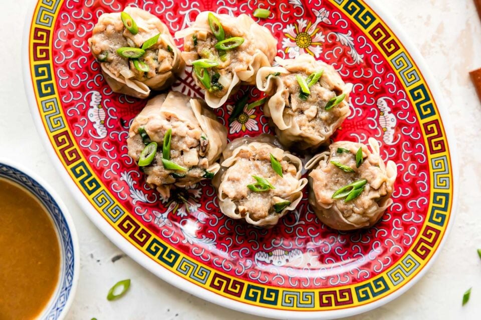 Six pork shumai dumplings rest atop a colorful platter that sits atop a creamy white textured surface. The pork shumai dumplings have been garnished with sliced green onions. A pair of wooden chopsticks rest alongside the platter and a small blue and white bowl filled with siu mai sauce rests alongside the platter.