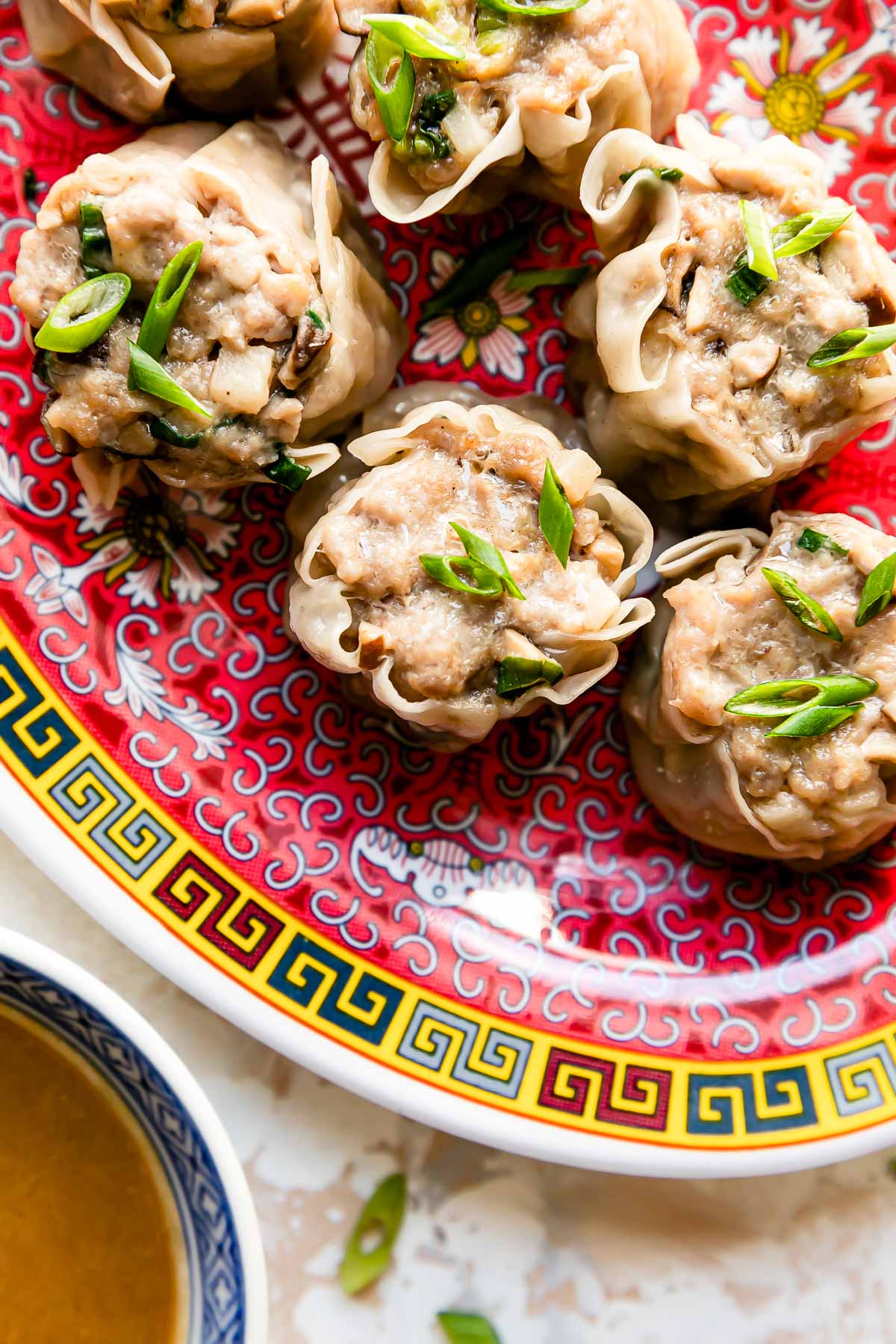 A close up shot of six pork shumai dumplings rest atop a colorful platter that sits atop a creamy white textured surface. The pork shumai dumplings have been garnished with sliced green onions. A pair of wooden chopsticks rest alongside the platter and a small blue and white bowl filled with siu mai sauce rests alongside the platter.