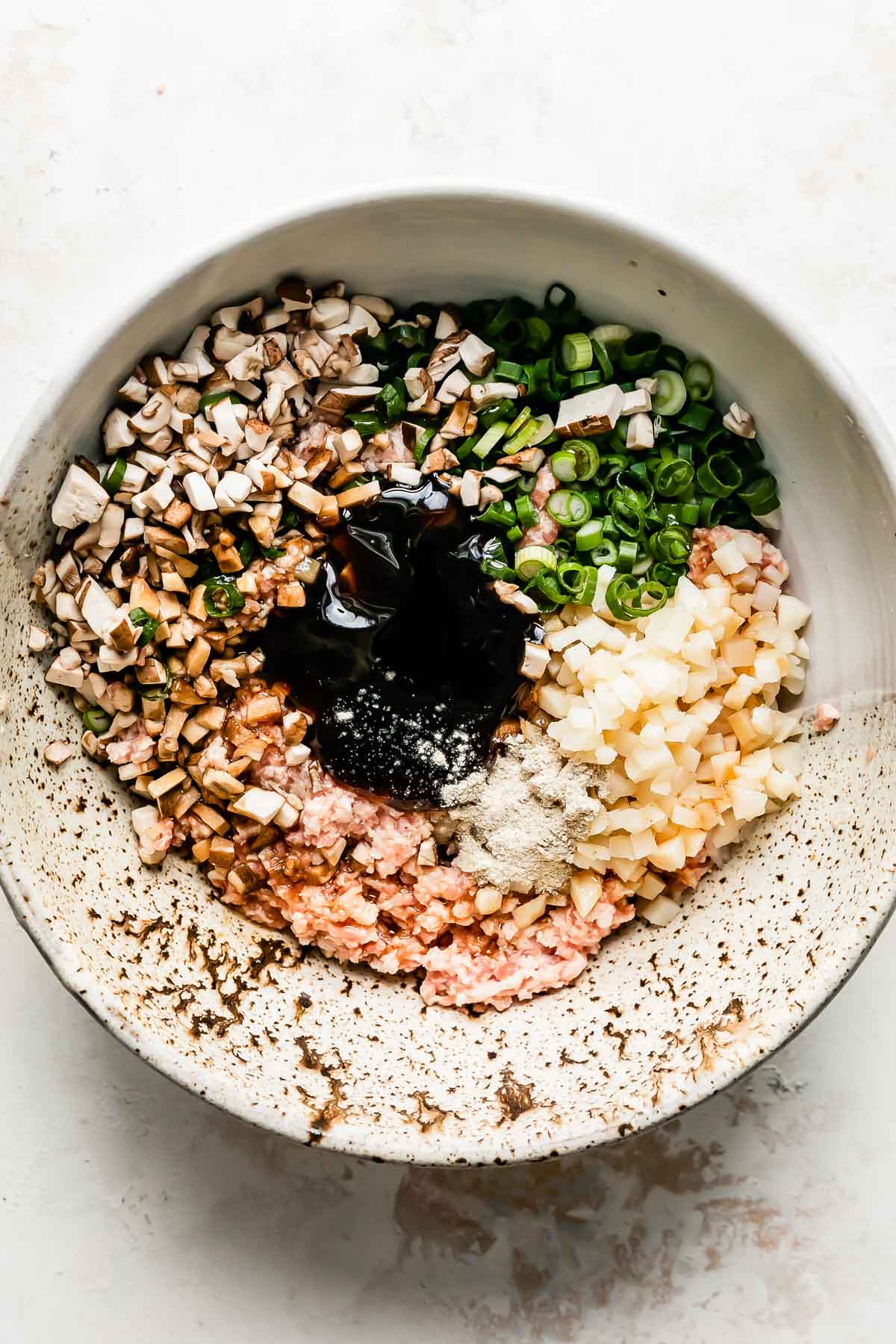 The components that make up pork shumai filling fill a large ceramic bowl that sits atop a creamy white textured surface: ground pork, cornstarch slurry, green onions, mushrooms, water chestnuts, oyster sauce, soy sauce, sugar, and white pepper.
