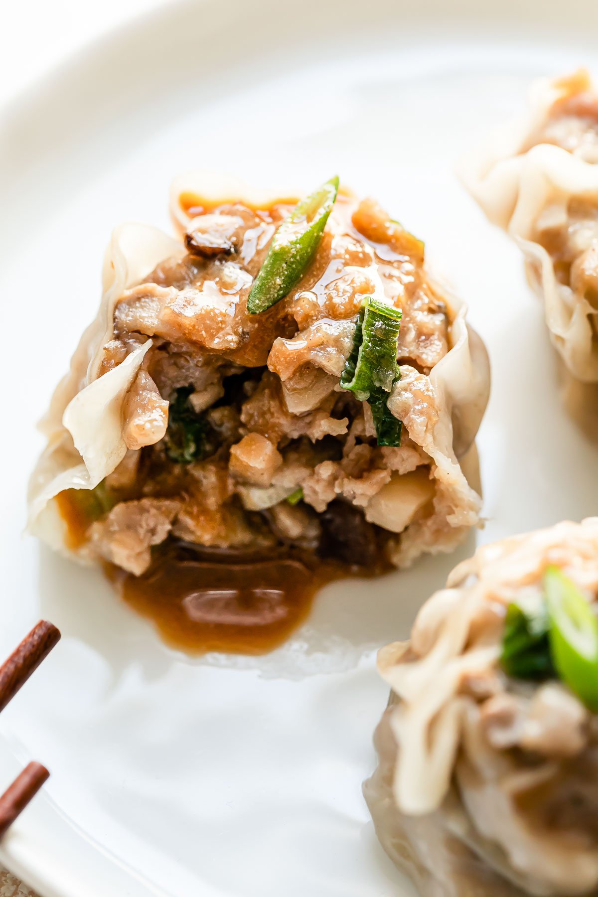 A close up macro shot of pork shumai served atop a small white plate. A single shumai dumpling has a bite taken out of it and has been drizzled with siu mai sauce. A pair of wooden chopsticks rests atop the small white plate.