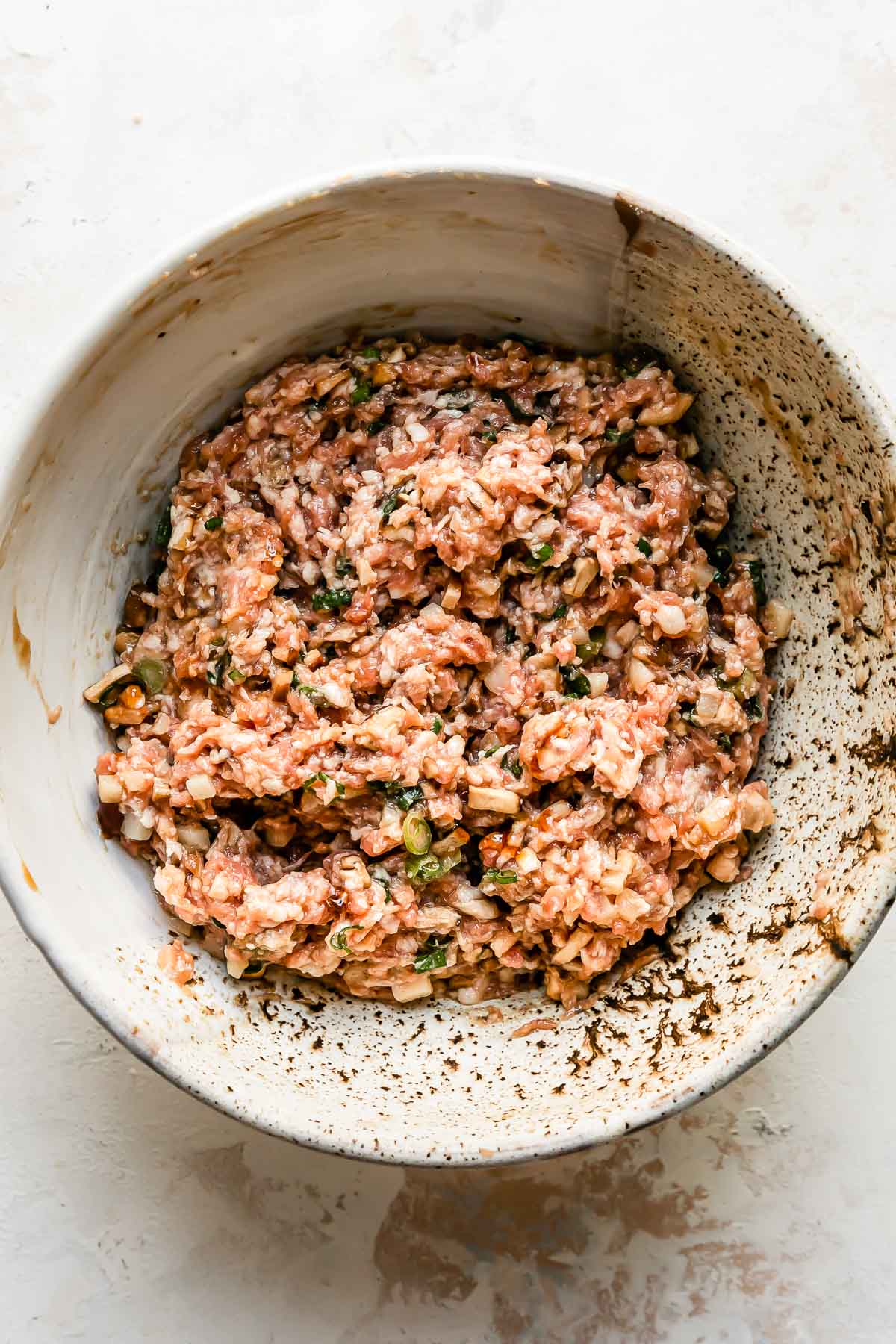 Shumai dumpling filling rests inside of a large ceramic bowl that sits atop a creamy white textured surface.