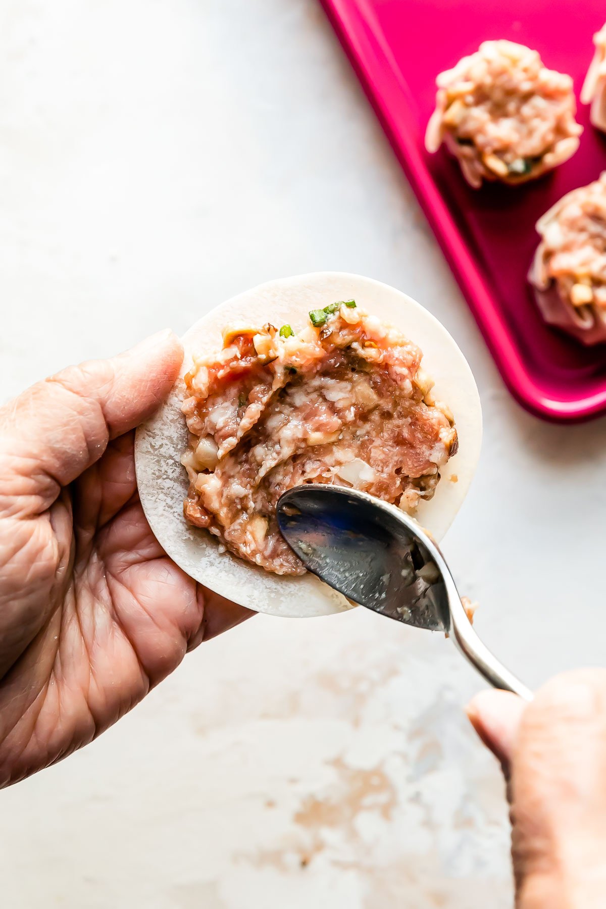 How to make a shumai dumpling, step 3: Siu mai assembly. A woman's hands spread pork shumai filling atop a won ton wrapper with the back of a spoon to begin assembling a shumai dumpling over a creamy white textured surface. A pink baking sheet filled with a few assembled shumai dumplings rests atop the surface.