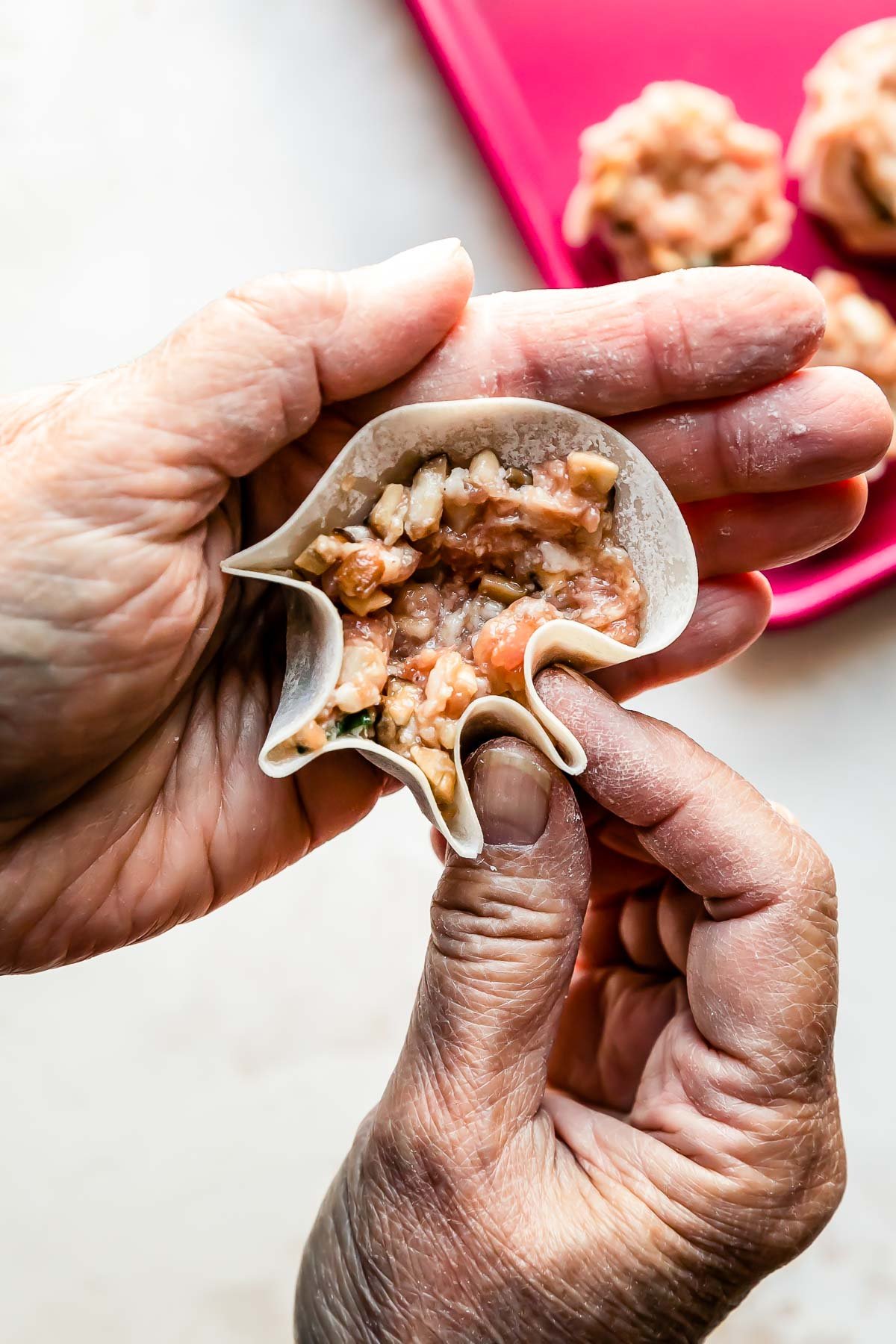 A woman's hands form a shumai dumpling into a purse shape over a creamy white textured surface. A pink baking sheet filled with a few assembled shumai dumplings rests atop the surface.