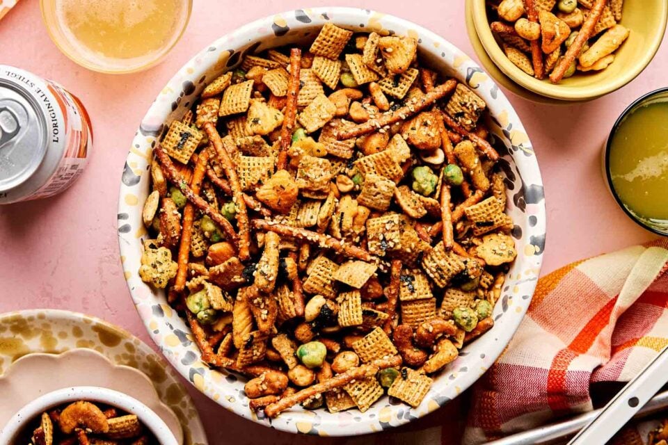 An overhead shot of a large patterned bowl of furikake chex mix on a light pink surface. The bowl is surrounded by small dishes of chex mix and a plaid cloth.