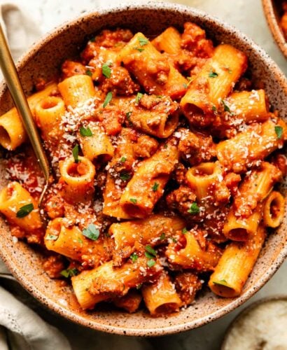 An overhead shot of a serving of sausage ragu pasta in a brown stoneware bowl sitting atop an off-white surface. A small bowl of grated parmesan and an off-white cloth sit beside the bowl.