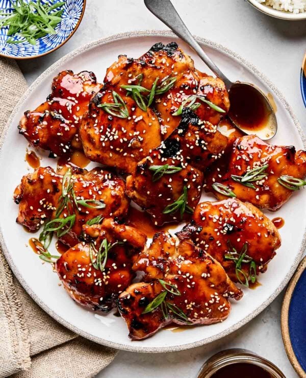 An overhead shot of teriyaki chicken garnished with sesame seeds and green onions on a white plate atop a white marbled surface. The plate is surrounded by dishes of rice, green onions, and teriyaki sauce.
