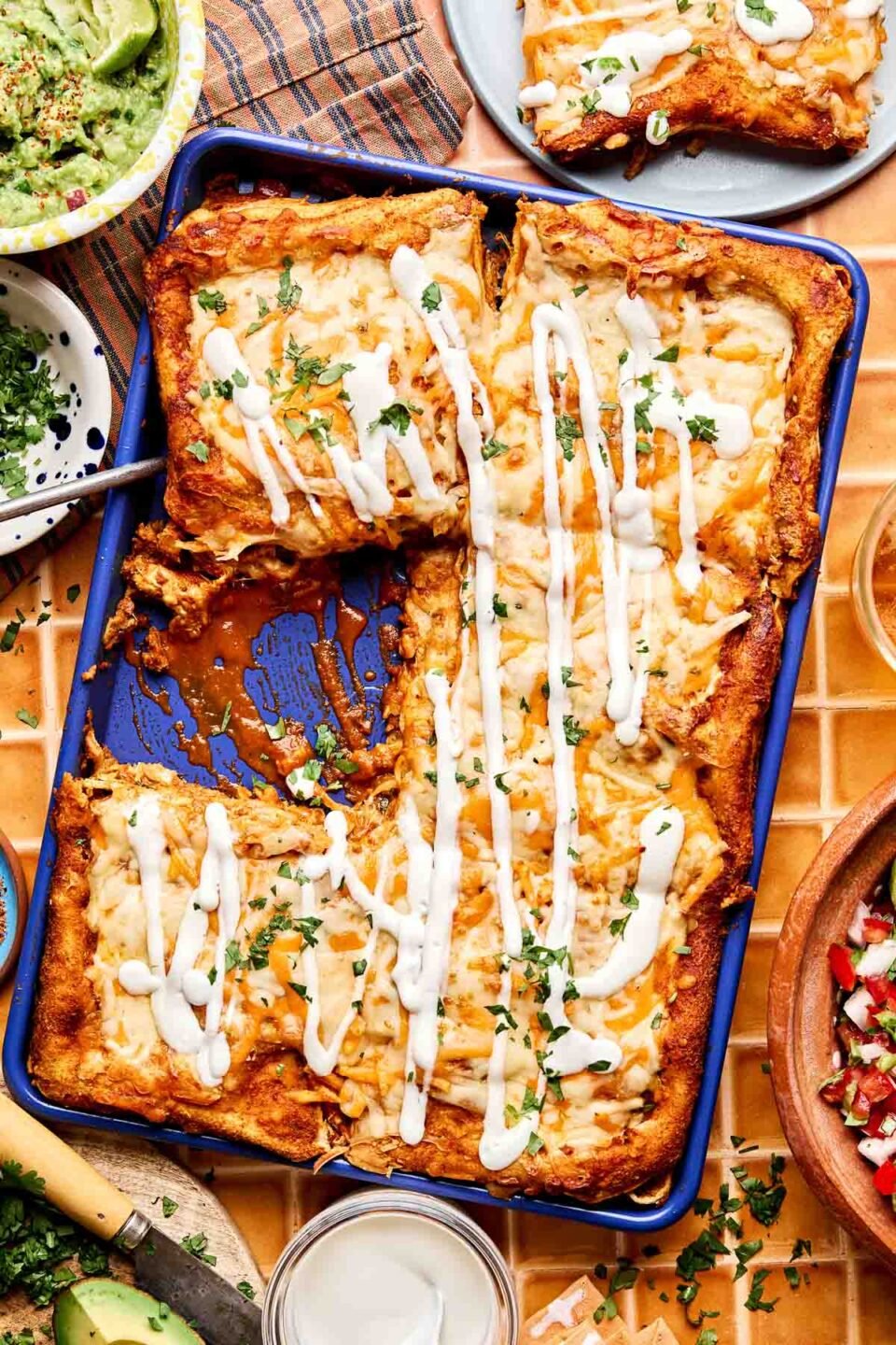 A blue baking dish filled with cheesy enchiladas topped with white sauce and chopped herbs, sitting on a tiled table surrounded by bowls of guacamole, pico de gallo, and avocado slices.