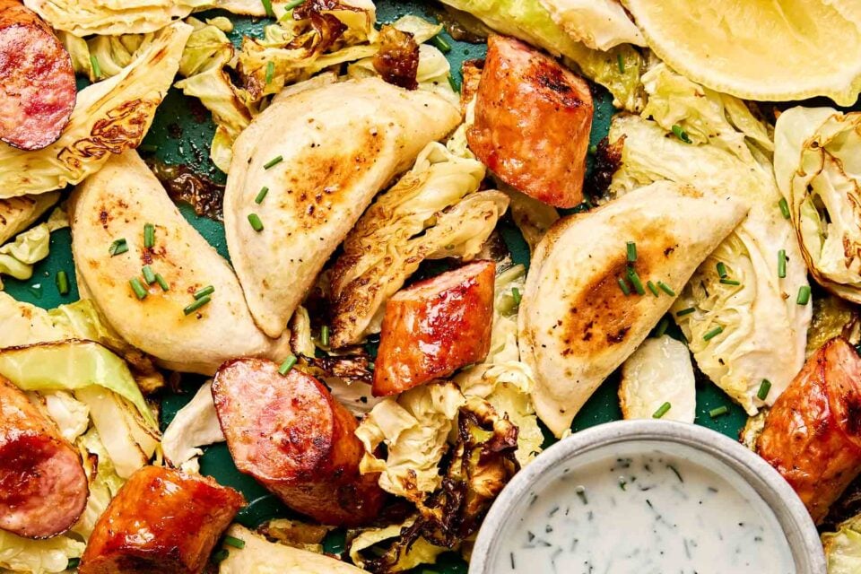 Golden-brown pierogies, sliced sausage, and roasted cabbage are arranged on a tray, garnished with chopped chives. A small bowl of creamy dipping sauce is placed in the corner.