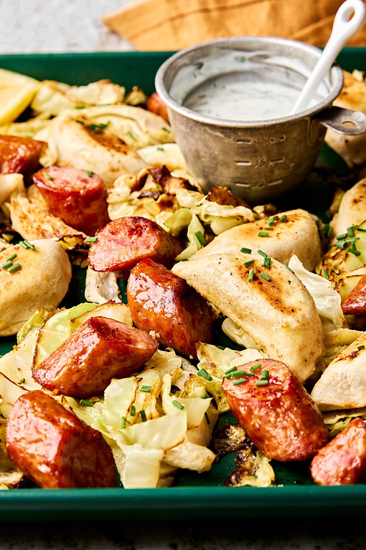 A green pan filled with roasted pierogies, sliced kielbasa, and cabbage, topped with chopped chives. A metal measuring cup of creamy dipping sauce with a spoon sits on the tray.