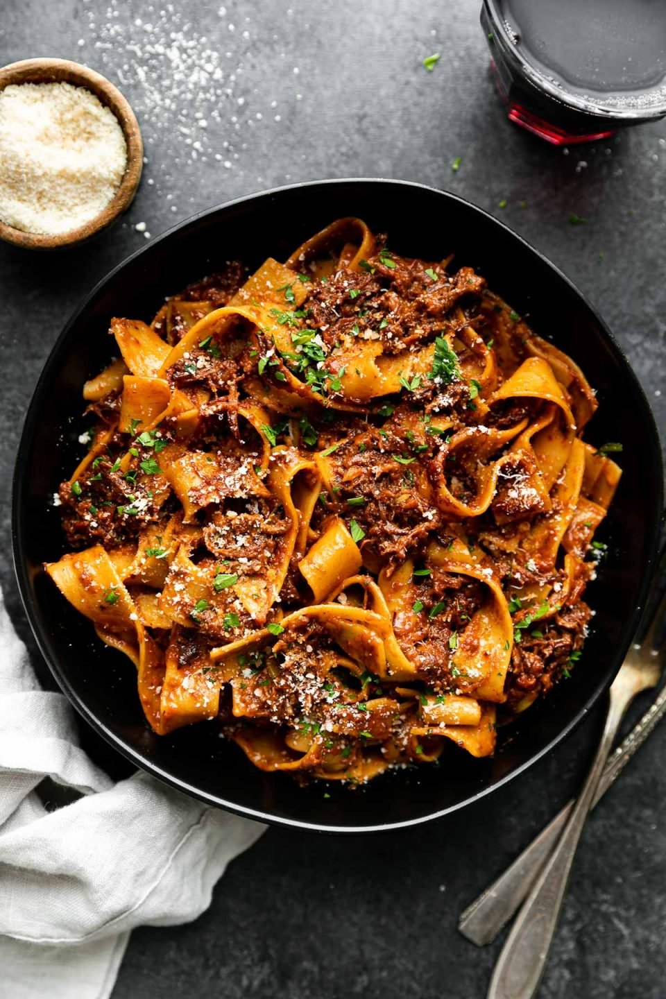 Braised Short Rib Ragu Pappardelle shown in a pasta bowl, topped with grated parmesan & fresh parsley. The bowl sits atop a black surface surrounded by a clear glass filled with red wine, a small wooden pinch bowl filled with grated parmesan, a gray linen napkin, & a set of silver silverware.