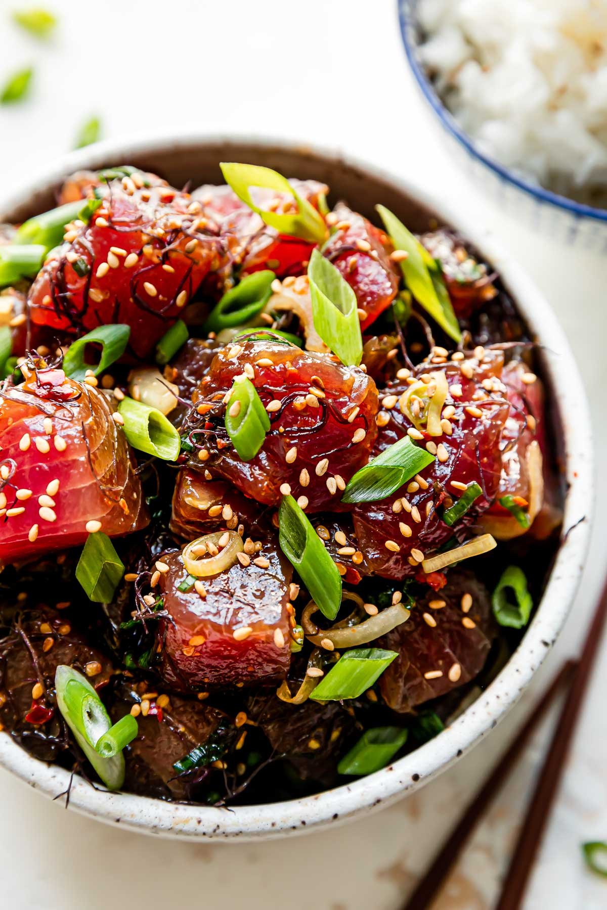 An overhead and close up macro shot of ahi poke inside of a small ceramic bowl that sits atop a creamy white textured surface. It is garnished with toasted sesame seeds and thinly sliced green onion. A small blue and white bowl filled with white rice and a pair of wooden chopsticks rests alongside the bowl of tuna poke at center.