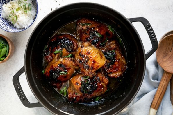 Shoyu chicken in a black dutch oven atop a white surface. The chicken is garnished with sliced green onions & sesame seeds. The dutch oven is surrounded by a blue linen napkin & serving spoons, & 2 small bowls, one filled with rice & the second with sliced green onions.