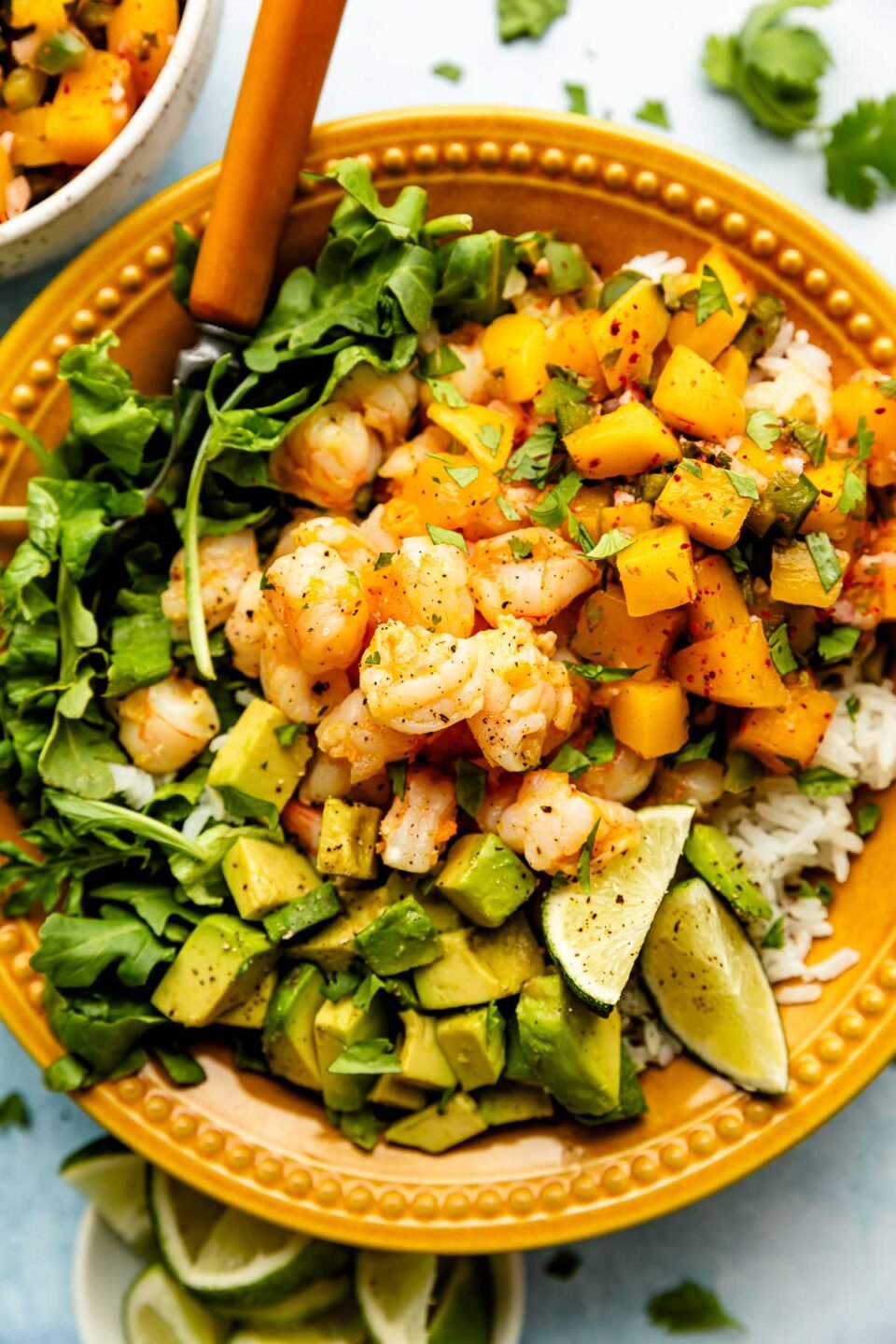 An overhead shot of an assembled shrimp bowl with lettuce, rice, avocado, and mango salsa in a light orange bowl atop a light blue surface. The bowl is surrounded by other assembled bowls and bowls of mango salsa, cilantro, and lime wedges.
