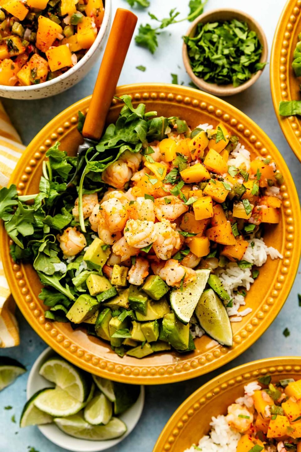 An overhead shot of an assembled shrimp bowl with lettuce, rice, avocado, and mango salsa in a light orange bowl atop a light blue surface. The bowl is surrounded by other assembled bowls and bowls of mango salsa, cilantro, and lime wedges.