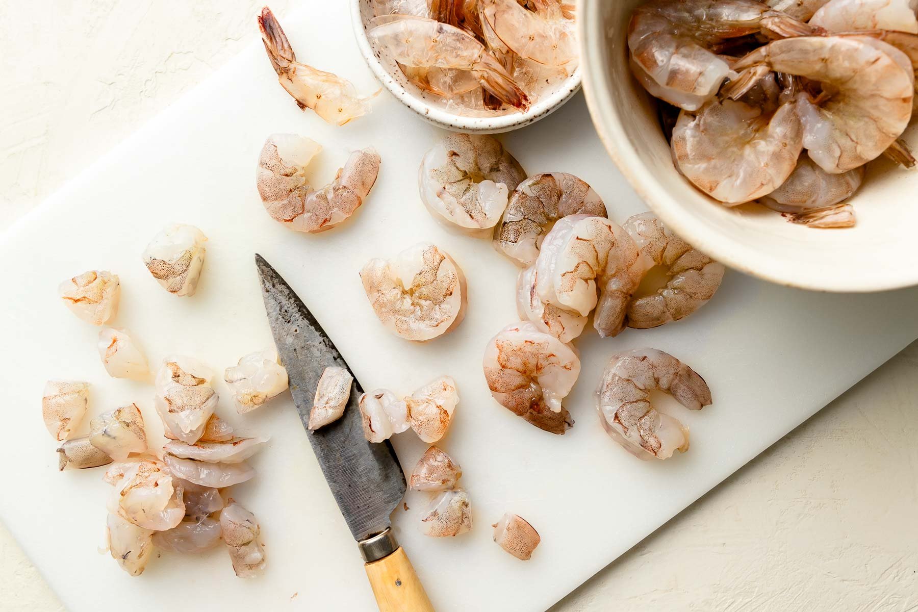 An overhead shot of raw shrimp being chopped into fine pieces on a white cutting board atop an off-white surface.