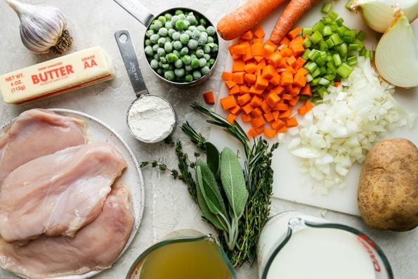 Boneless, skinless chicken breasts, yellow onion, carrots, celery, Russet potato, garlic, all-purpose flour, chicken stock, whole milk, fresh rosemary, fresh thyme, fresh sage, frozen peas arranged on a creamy white textured surface.