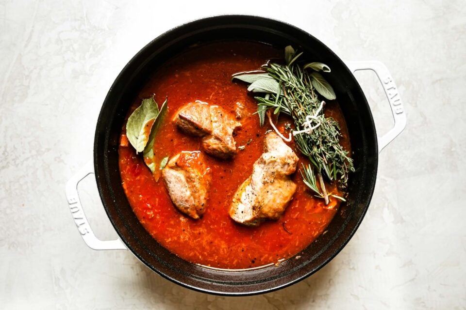 Browned pork shoulder with bay leaves & fresh herbs, nestled in tomato sauce in a large white Dutch oven.