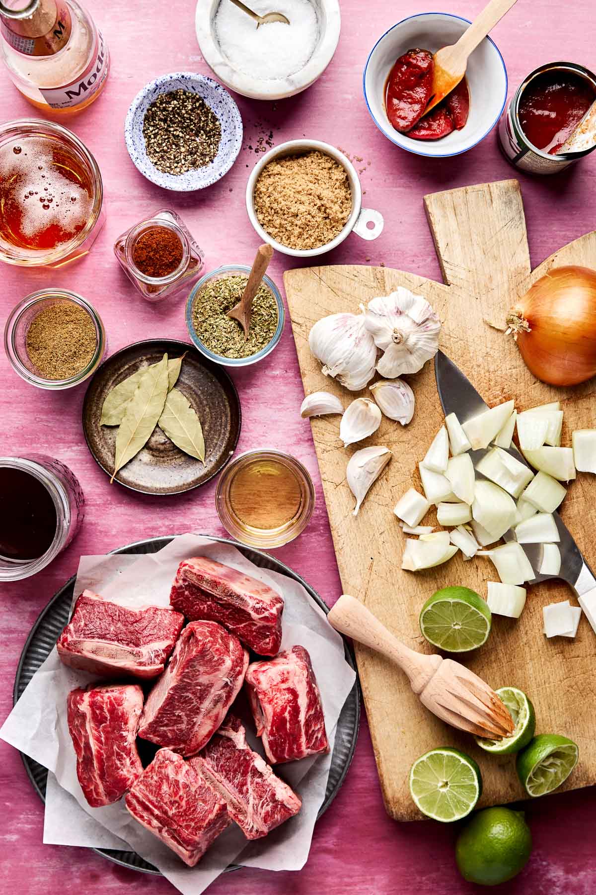 An overhead shot of ingredients displayed in various dishes and on a wooden board atop a pink surface: raw short ribs, limes, yellow onion, garlic, beer, vegetable oil, salt and pepper, light brown sugar, chipotle peppers, chili powder, ground cumin, dried oregano, bay leaves, and beef broth.