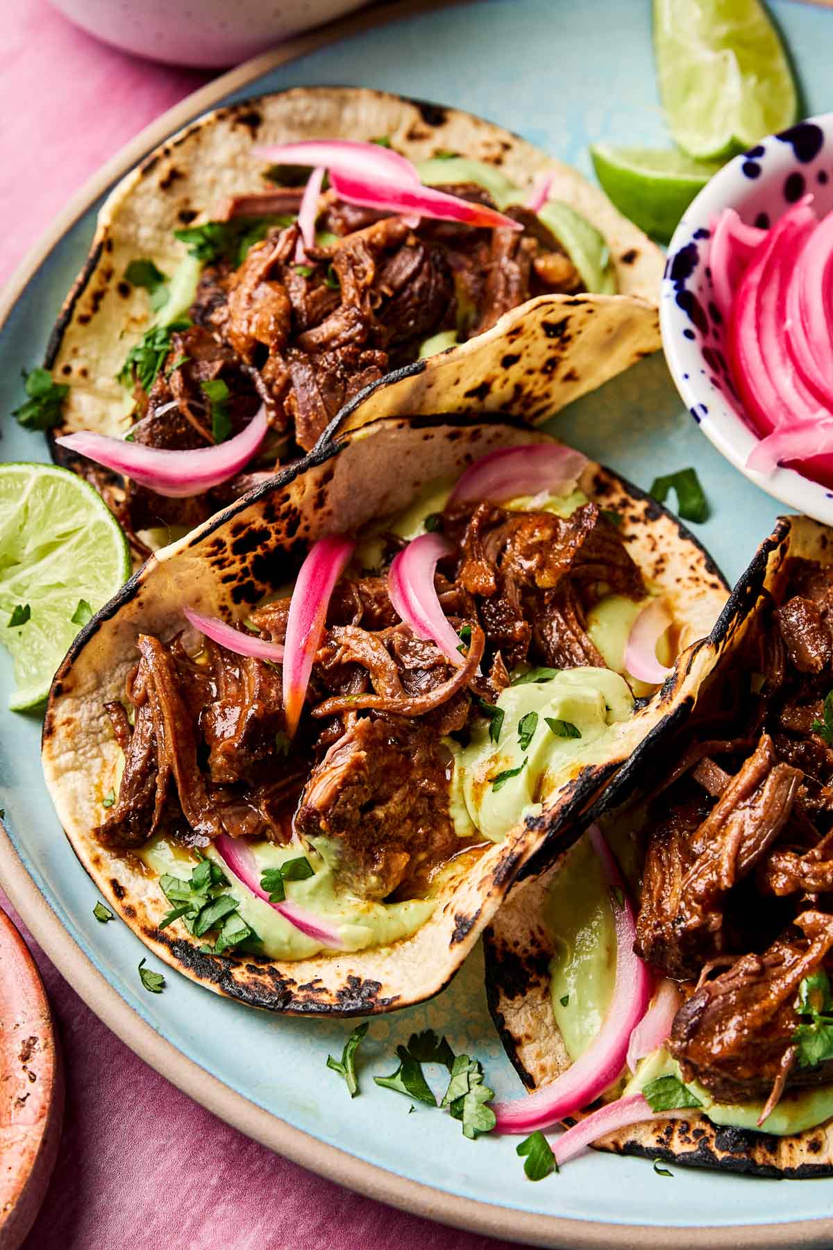 An overhead shot of a light blue plate holding three braised short rib tacos garnished with pickled red onions and fresh cilantro on a pink surface.
