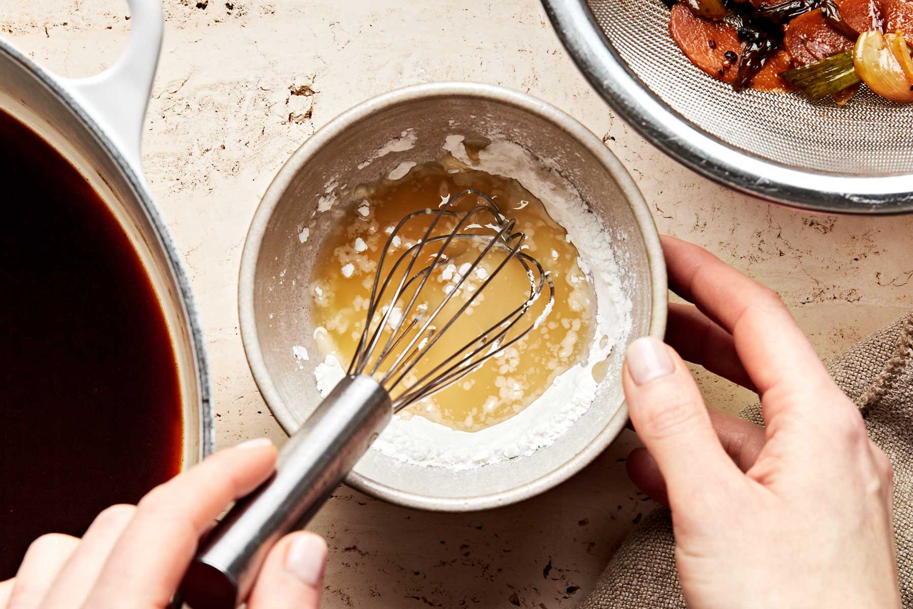 An overhead shot of a woman's hands mixing a cornstarch slurry in a beige stoneware bowl atop a textured beige surface. A strainer with strained aromatics and the pot of braising liquid sit alongside the bowl.
