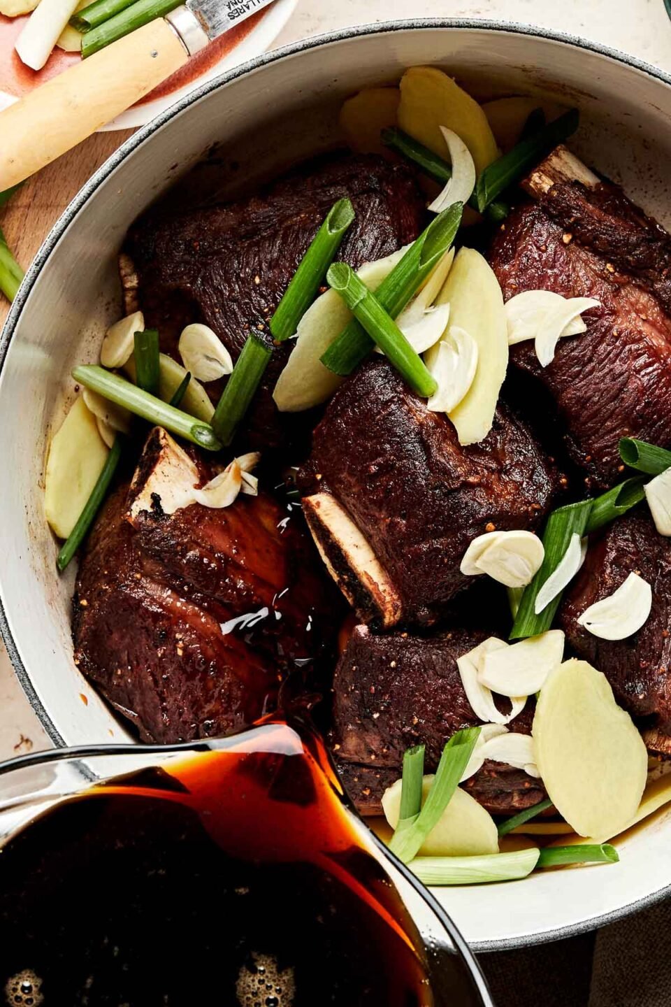 An overhead shot of six browned short ribs in a white pot with ginger, garlic, and green onions. Braising liquid is being poured into the pot from a liquid glass measuring cup. The pot sits on an off white surface alongside a beige cloth, a wooden cutting board, and extra ginger, green onions, and garlic.