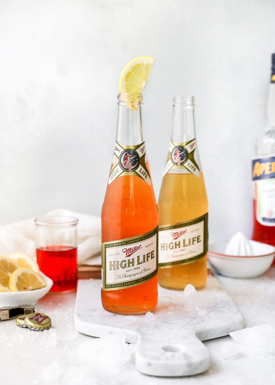 2 Spaghett Cocktails served in Miller High Life bottles. The beer bottles are on a white surface, surrounded by lemon wedges, ice and some Aperol liquor.