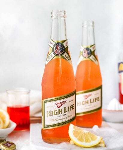2 Spaghett Cocktails served in Miller High Life bottles. The beer bottles are on a white surface, surrounded by lemon wedges, ice & some Aperol liquor.