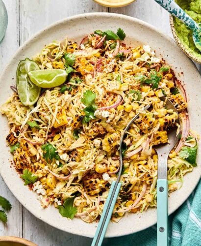A plate of Mexican street corn salad with grilled corn, shredded cheese, red onion, cilantro, and lime wedges, served with blue-handled tongs on a rustic white surface. Small bowls with toppings are nearby.