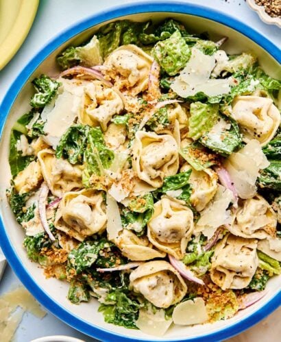 A bowl of tortellini Caesar salad with cheese tortellini, romaine lettuce, shaved parmesan, red onion, croutons, and Caesar dressing, garnished with cracked black pepper.