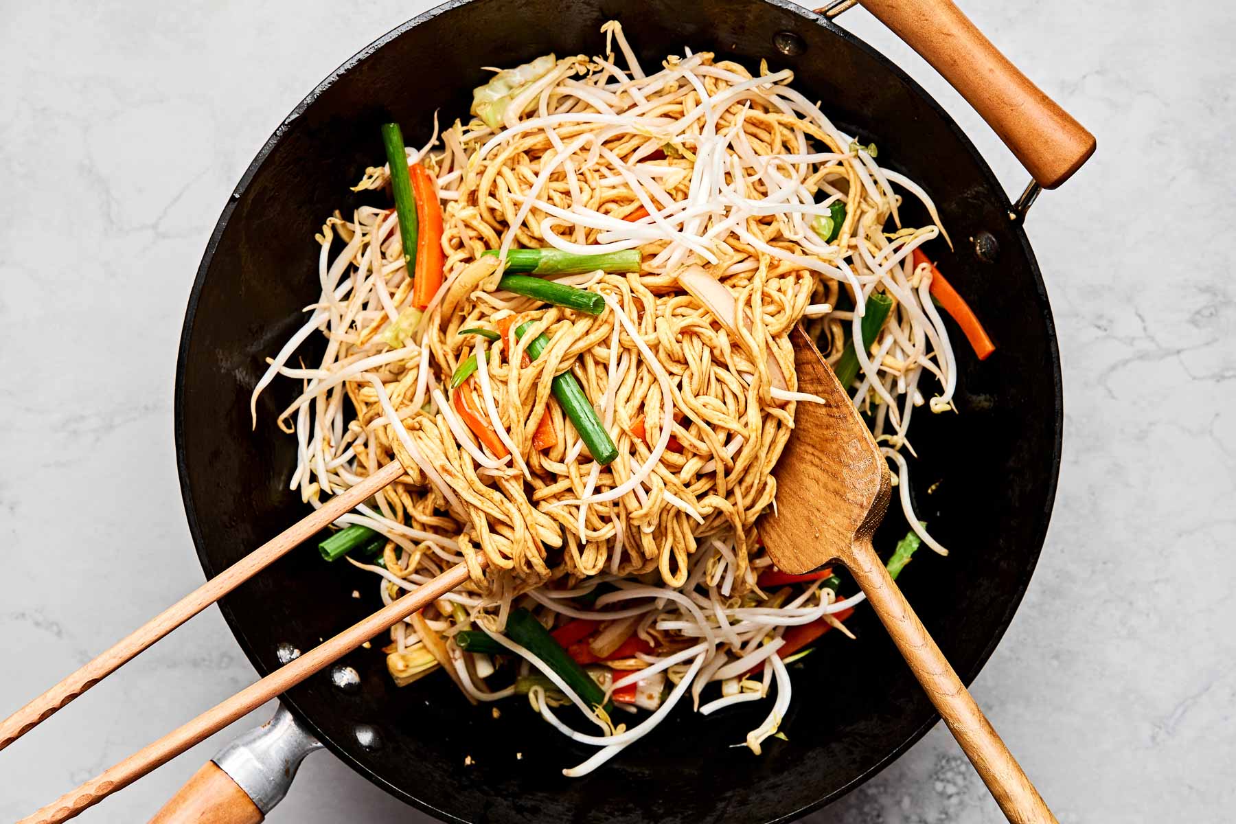 An overhead shot of bean sprouts being tossed with noodles and veggies in a large black wok on a white marbled surface.