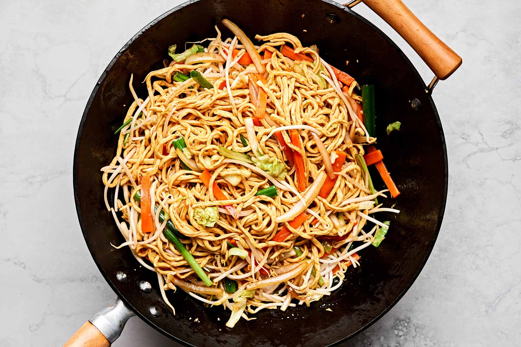 An overhead shot of vegetable stir fried noodles in a large black wok on a white marbled surface.