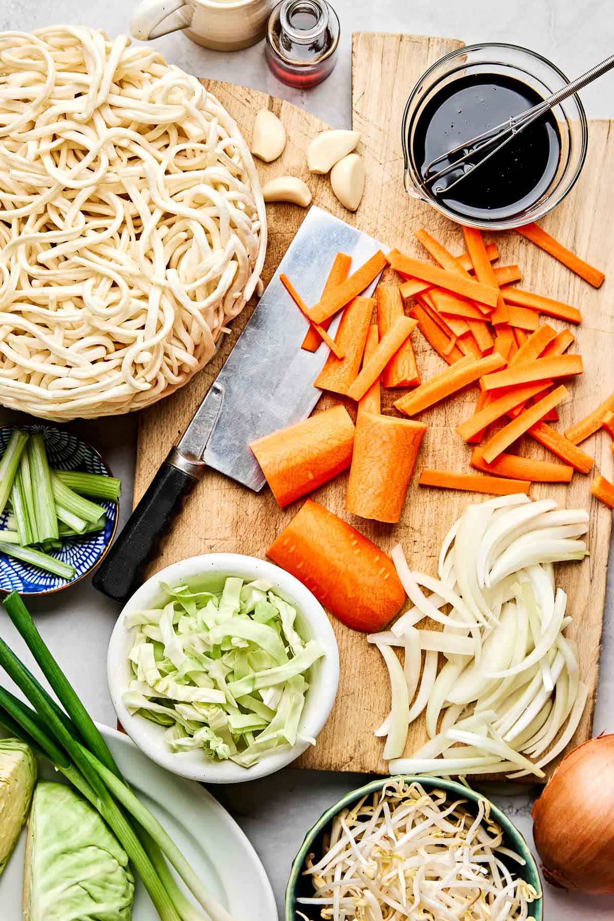An overhead shot of ingredients displayed on a white marbled surface: yee mein noodles, sliced carrots, yellow onion, garlic, green onions, cabbage, bean sprouts, soy sauce, shoyu, and oyster sauce.
