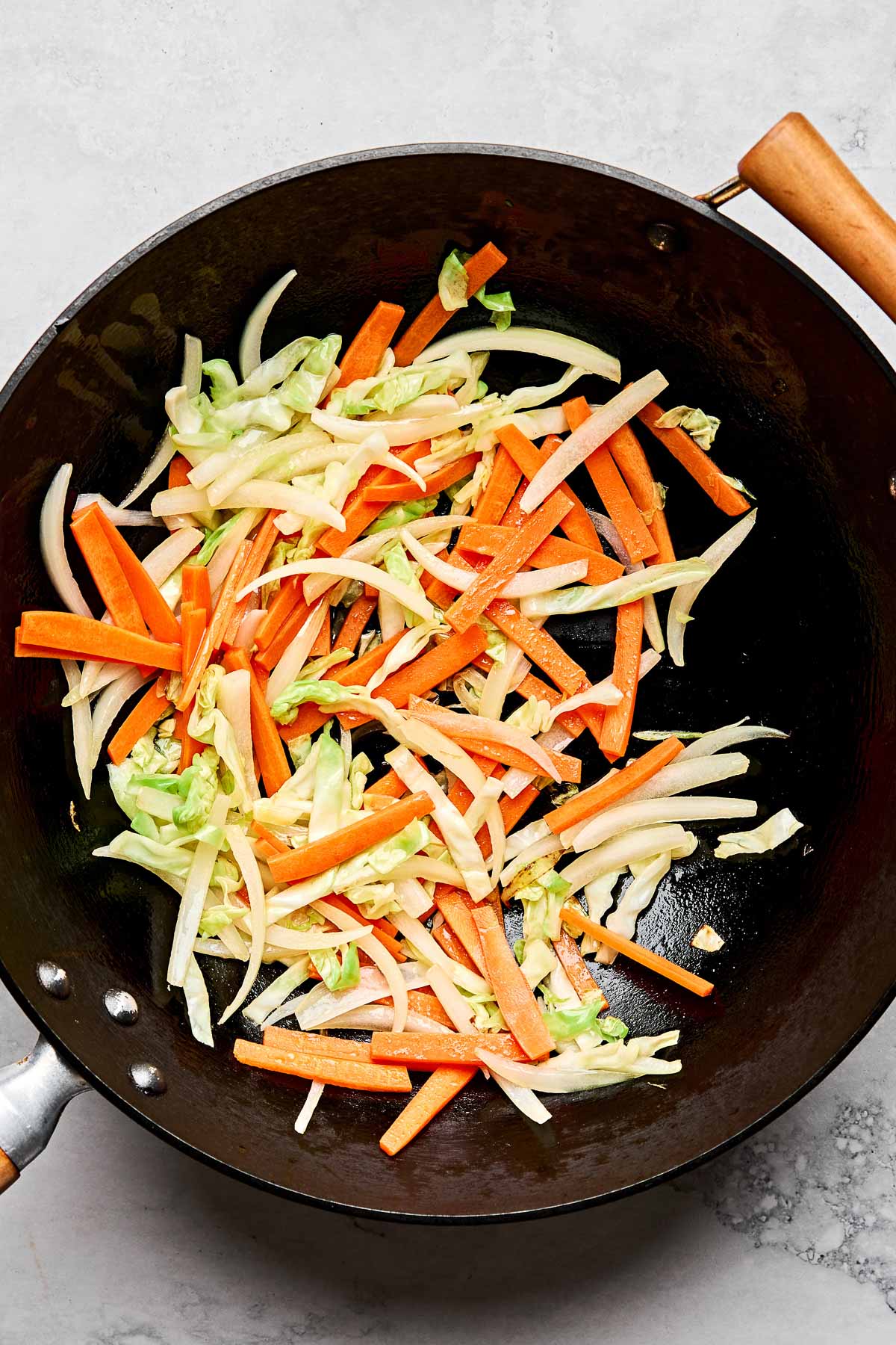 An overhead shot of julienned carrots, onion, and cabbage in a large black wok on a white marbled surface.