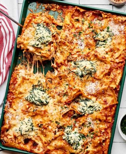 A baking sheet filled with cheesy baked lasagna, cut into squares. Some pieces are being served, showing stretchy melted cheese, tomato sauce, and dollops of creamy spinach ricotta on top. A red and white towel is beside the tray.