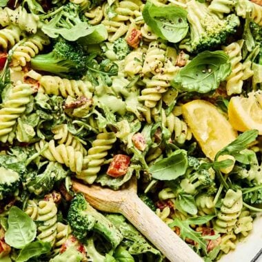 A close-up overhead shot of creamy vegan pasta in a white skillet, topped with basil leaves and lemon wedges.